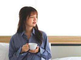 asiatisk ung kvinna Sammanträde på säng innehav kopp av kaffe, ser bort. foto