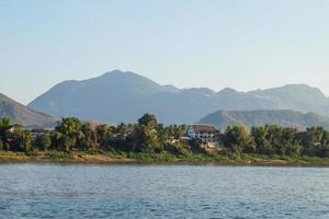 se på flodstrand med palmer träd, hus av lokalbefolkningen och bergen på bakgrund. mekong flod, luang prabang, laos. foto