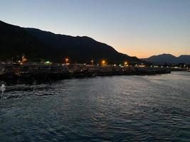 skön kust och strand med konturer av bergen mot de bakgrund av solnedgång från de hav eller hav och brinnande lampor av de stad och hamn med fartyg foto