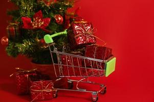en handla vagn full av gåvor mot röd bakgrund nära små jul träd. jul eller ny år handla foto
