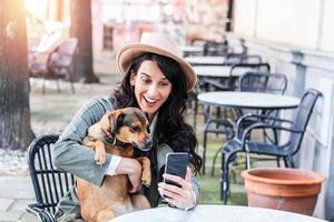 ung kvinna med henne hund i sällskapsdjur vänlig cafeteria. flicka dricka coffe på lokal- kaffe affär med henne sällskapsdjur hund tar en selfie med mobil telefon foto