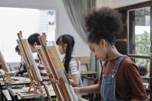 afrikansk amerikan flicka koncentrat på akryl Färg bild målning på duk med studenter grupp i konst klassrum, kreativ inlärning med talanger och Kompetens i elementärt skola studio utbildning. foto