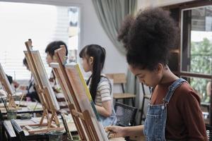 afrikansk amerikan flicka koncentrat på akryl Färg bild målning på duk med studenter grupp i konst klassrum, kreativ inlärning med talanger och Kompetens i elementärt skola studio utbildning. foto