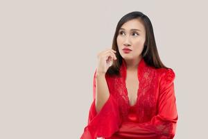 bild av tänkande sexig lady stående isolerat över rosa bakgrund. ser bort. de hand nära de ansikte och mun. asiatisk kvinna i en röd satin nattkläder bär spets morgonrock tänkande utseende vänster. foto