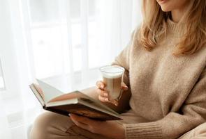 stänga upp kvinnors händer och fingrar med en skön franska manikyr innehav kopp transparent glas med kaffe med vit skum.kopia Plats för text, baner. beige stickat Tröja och bakgrund. foto