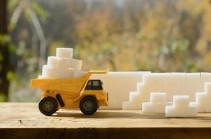 en små gul leksak lastbil är lastad med vit socker kuber nära de socker vägg. en bil på en trä- yta mot en bakgrund av höst skog. extraktion och transport av socker foto