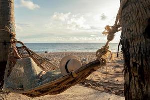 skön ung asiatisk kvinna med hatt avkopplande avkopplande i en hängmatta mellan handflatan träd, sommar Lycklig strand semester begrepp. foto