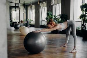 smal kvinna som sträcker sig tillbaka med träningsboll medan du tränar i modern fitnessstudio foto