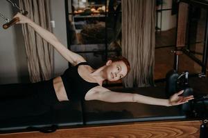 ung kvinnlig pilatesinstruktör liggande i sträckande armposition på cadillac reformer foto