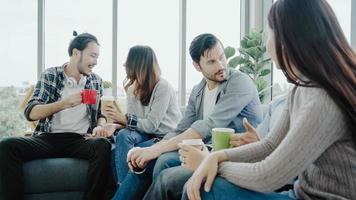 mångfald av ungdomars grupplag som håller kaffekoppar och diskuterar något med leende när man sitter i soffan på kontoret. kaffepaus på kreativt kontor. foto