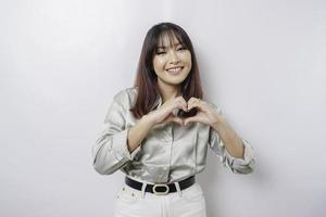 ett attraktiv ung asiatisk kvinna bär en salvia grön skjorta känner Lycklig och en romantisk former hjärta gest uttrycker anbud känslor foto