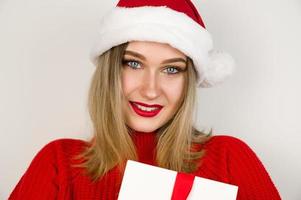 närbild porträtt av kvinna blond i röd Tröja med santa hatt leende och innehav närvarande. chistmas afton, helgdagar begrepp foto