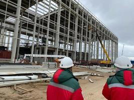 tillbaka skott av arbetstagare entreprenör bär hård hatt och säkerhet västar promenader på industriell byggnad konstruktion webbplats. i de bakgrund kran, skyskrapa betong formning ramar foto