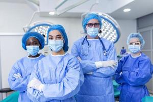 multietnisk grupp av fyra sjukvård arbetare, en team av läkare, kirurger och sjuksköterskor, utför kirurgi på en patient i en sjukhus rörelse rum. de är ser på de kamera. foto