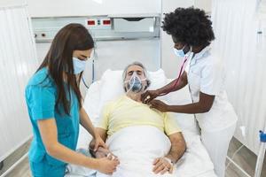 ung läkare och sjuksköterska bär en kirurgisk mask kontroll på en senior manlig patient bär en positiv tryck syre mask till hjälpa andas i en sjukhus säng under de covid-19 pandemi foto