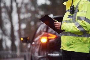 manlig polis officer i grön enhetlig stående med anteckningsblock nära bil foto