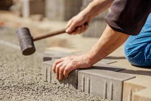 ung man om grå betong stenläggning plattor i hus gård på grus fundament bas. bemästra lägger stenläggning stenar. trädgård tegel väg stenläggning förbi professionell utläggare arbetstagare. reparation trottoar. foto