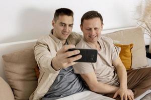 Lycklig Gay par med tillfällig kläder utgifterna tid tillsammans på Hem och framställning selfie på smartphone. homosexuell relationer och alternativ kärlek. mysigt interiör. foto