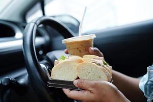asiatisk kvinna förare håll is kaffe kopp och smörgås bröd för äta och dryck i bil, farlig och risk ett olycka. foto