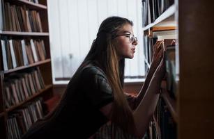 sökande för rätt information. mot fönster. kvinna studerande är i bibliotek den där full av böcker. uppfattning av utbildning foto