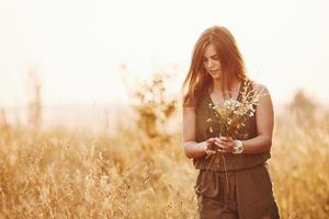 beautful flicka promenader genom de fält med hög gräs och samlar blommor. Fantastisk solljus foto