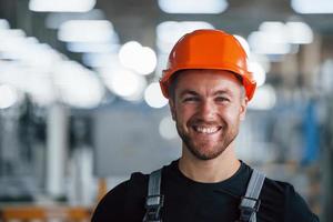 leende och Lycklig anställd. porträtt av industriell arbetstagare inomhus i fabrik. ung tekniker med orange hård hatt foto