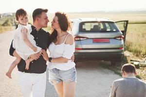 fotograf sitter ner. familj ha några Bra tid på landsbygden nära silver- bil på solnedgång foto