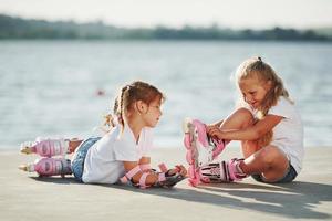 två liten flickor med vält skridskor utomhus nära de sjö på bakgrund foto