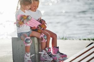 två liten flickor med vält skridskor utomhus nära de sjö på bakgrund foto