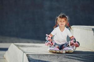 söt liten flicka med vält skridskor utomhus sitter på de ramp för extrem sporter foto