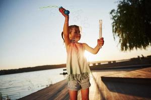 kväll tid. Lycklig liten flicka spelar med bubblor nära de sjö på parkera foto