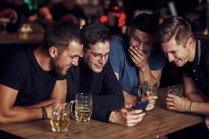 rolig innehåll på smartphone. tre vänner vilar i de pub med öl i händer. har konversation foto