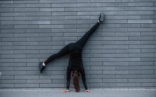 ung sportigt flicka i svart sportkläder håller på med hård stå på händer övningar utomhus nära grå vägg foto
