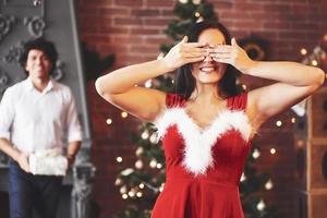 skön kläder. kvinna i röd klänning beläggning henne ögon och kommer motta jul gåva från pojkvän foto