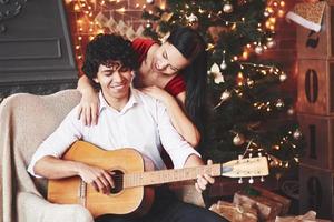 innehar akustisk gitarr. skön par fira ny år i de dekorerad rum med jul träd Bakom foto