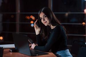 stor smartphone nära de öra. attraktiv brunett affärskvinna Arbetar ensam i de kontor på nightime foto