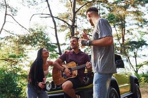 sång låtar tillsammans. vänner ha trevlig helgen utomhus nära deras grön bil med akustisk gitarr foto