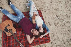 akustisk gitarr nära dem. ung par ha picknick på de strand. liggande på de röd färgad filt foto