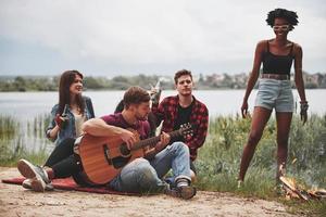 känna de musik. grupp av människor ha picknick på de strand. vänner ha roligt på helgen tid foto