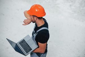 antenn se. med bärbar dator i händer. industriell arbetstagare inomhus i fabrik. ung tekniker med orange hård hatt foto