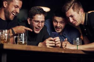rolig innehåll på smartphone. tre vänner vilar i de pub med öl i händer. har konversation foto