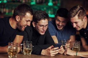 rolig innehåll på smartphone. tre vänner vilar i de pub med öl i händer. har konversation foto