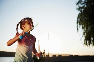 kväll tid. Lycklig liten flicka spelar med bubblor nära de sjö på parkera foto