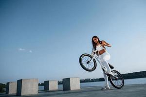 Foto i rörelse. håller på med stunts. kvinna ryttare är på de cykel på dagtid nära de sjö. kondition kvinna i sportigt kläder