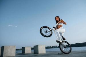 Foto i rörelse. håller på med stunts. kvinna ryttare är på de cykel på dagtid nära de sjö. kondition kvinna i sportigt kläder
