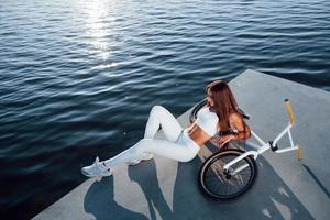 nära cykeln. fitness kvinna som vilar nära sjön på dagtid. vackert solljus foto