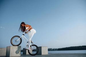 Foto i rörelse. håller på med stunts. kvinna ryttare är på de cykel på dagtid nära de sjö. kondition kvinna i sportigt kläder