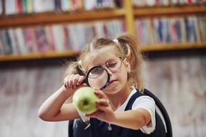 innehar förstoringsglas. söt liten flicka med pigtails är i de bibliotek. äpple på de böcker foto