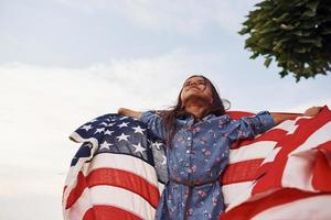 skön grön träd. patriotisk kvinna unge med amerikan flagga i händer. mot molnig himmel foto