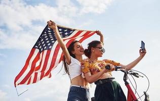 två patriotisk glad kvinnor med cykel och USA flagga i händer gör selfie foto